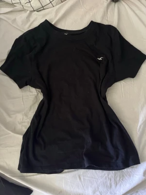 Svart t-shirt med liten logga - En klassisk svart t-shirt i bomull med rund hals och korta ärmar. På bröstet finns en liten vit logga som ger en clean och stilren look. Perfekt till jeans eller shorts för en enkel och snygg outfit.