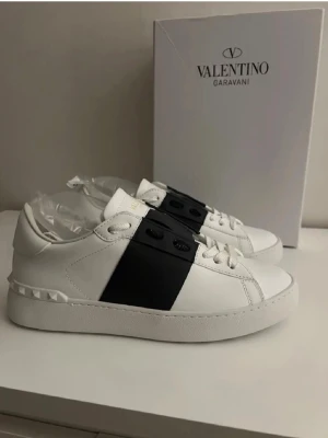 Valentino Garavani vita sneakers - Stilrena vita sneakers från Valentino Garavani med en bred svart rem över mitten och dekorativa nitar vid hälen. Skorna har en chunky vit sula med mönstrad undersida och är tillverkade i läder. Perfekta för dig som gillar exklusiv och modern design.