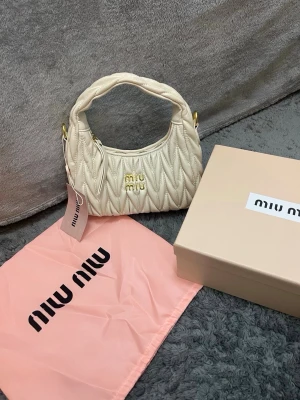 Vit quiltad handväska från Miu Miu - Supersnygg vit handväska från Miu Miu i quiltat läder med guldfärgade detaljer och logga framtill. Väskan har ett vadderat handtag och dragkedja upptill. Kommer med dustbag och originalkartong. Perfekt accessoar för att lyfta din outfit.