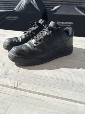 Nike Air Force 1 Low helsvart - Klassiska Nike Air Force 1 Low sneakers i helsvart med svart sula och svarta skosnören. Ikonisk Swoosh på sidan och metallbricka med AF1 på snörningen. Perfekta för en clean och stilren look.