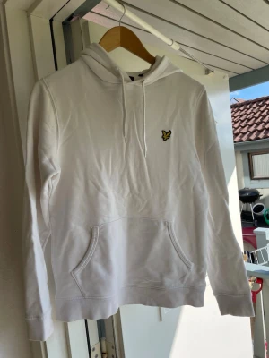 Vit hoodie från lyle&scott - Snygg vit hoodie med huva och dragsnören samt en broderad Lyle&scott logga på bröstet. Klassisk känguruficka framtill och ribbade muddar vid ärmslut och nederkant. Perfekt för en chill och stilren look.