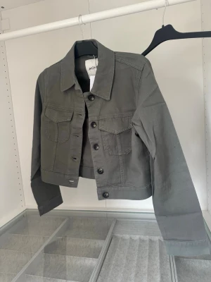 Aldrig använd Grå/grön kort jeansjacka från Monki - Snygg grå jeansjacka från Monki med kortare passform. Jackan har klassisk krage, två bröstfickor med lock och knappar framtill. Perfekt att slänga över en hoodie eller t-shirt för en chill look. Aldrig använd 