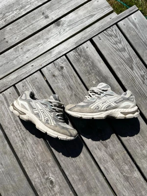 Asics gel nyc  - Säljer ett par grå sneakers med vita och silverfärgade detaljer. Skorna har meshpaneler för extra ventilation och reflekterande partier på sidorna. Klassisk snörning och robust sula för bra komfort. Perfekta för dig som gillar sportig stil.