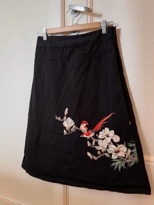Svart blommig kjol från KappAhl - Svart långkjol från KappAhl med ett snyggt blommigt tryck och en röd fågel på ena sidan. Kjolen har en rak passform och är gjord i mjukt bomullstyg. Perfekt för dig som vill ha något unikt och stilrent i garderoben.