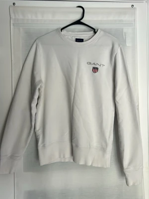 Vit sweatshirt från GANT - Klassisk vit sweatshirt från GANT med rund halsringning och broderad logga samt sköld på bröstet. Tröjan har långa ärmar och ribbade muddar vid ärmslut och nederkant. Perfekt för en clean och stilren look.