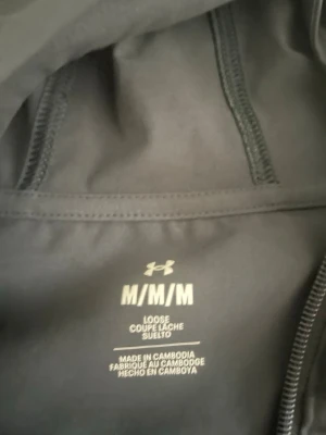 Svart träningsjacka från Under Armour - Säljer en svart träningsjacka från Under Armour i storlek M. Jackan har lös passform, dragkedja och elastiska muddar. Under Armour-logga på bröstet och text längs ena ärmen. Perfekt för träning eller som sportig vardagsstil. Priset kan diskuteras 