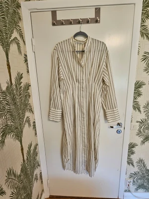 Midiklänning Sabosse dress - Midiklänning i vit och beige med tunna svarta ränder. Klänningen har lång ärm, ståkrage och knäppning framtill. Perfekt för en avslappnad look. Använd ett fåtal gånger. Nypris 2 300 kr. 