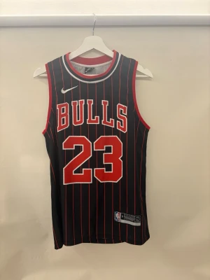 Chicago Bulls basketlinne #23 Jordan - Snyggt basketlinne från Nike med Chicago Bulls tryck och nummer 23, Jordan. Svart med röda och vita ränder, röda detaljer vid ärmhål och hals. Stort tryck fram och bak, meshmaterial som andas. Perfekt för basket eller streetwear.