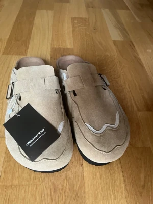 Beige mockatofflor med spänne - Stilrena beige tofflor i mocka från decarbo med dekorativa sömmar och justerbart spänne i silver på sidan. Tofflorna har öppen häl och svart sula, vilket ger en avslappnad men trendig look. Perfekta för dig som gillar minimalistisk design och mjuka material.