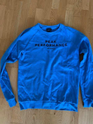 Blå sweatshirt från Peak Performance - Snygg blå sweatshirt från Peak Performance med svart broderad logga på bröstet. Tröjan har rund halsringning, långa ärmar och ribbade muddar vid ärmslut och nederkant. Perfekt för en avslappnad och sportig stil.