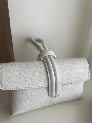 Vit axelväska i skinnimitation - Snygg vit axelväska i skinnimitation med unik knutdetalj framtill. Väskan har justerbar axelrem och stilren, rektangulär form. Perfekt för dig som gillar minimalistisk design och vill ha en väska som sticker ut lite extra. Använd 1 gång. Köpte på Åhléns för 1000kr