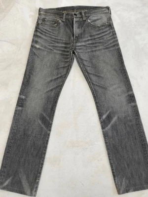 Grå raka jeans med slitningar - Säljer ett par grå jeans med raka ben och snygga slitningar framtill och baktill. Jeansen har klassisk femficksmodell och en läderpatch bak i midjan. Materialet är denim och färgen är tvättad grå med ljusare partier för en cool look.