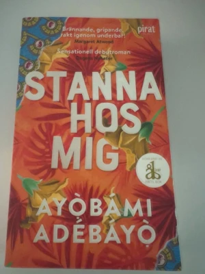 Stanna hos mig av Ayòbámi Adébáyò - Färgstark pocketbok med röd och orange omslag, dekorerad med blommor och blad. Romanen är skriven av Ayòbámi Adébáyò och utgiven av Piratförlaget. Boken handlar om kärlek, familj och traditioner i Nigeria under 80-talet.
