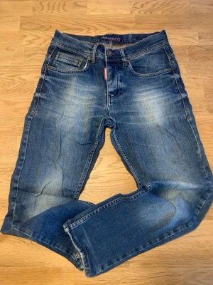 Blå jeans med ICON-lapp DSQUARED2 - Säljer ett par snygga blå jeans Dsquared2 med slitningar och tvättade detaljer. Klassisk femficksmodell och läderlapp med texten ICON bak i midjan. Jeansen har raka ben och normal midja. Perfekta för en avslappnad och trendig look.
