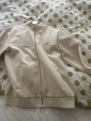 Beige zip hoodie från Les Deux - Snygg beige hoodie från Les Deux med dragkedja framtill och två fickor. Tillverkad i ett mjukt material som känns skönt mot huden. Minimalistisk design med diskret logga på bröstet. Perfekt för en clean och stilren look.