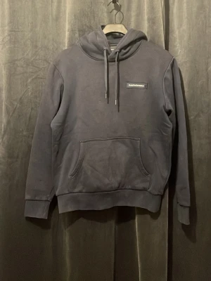 Mörkgrå Peak Performance hoodie med snörning - Säljer en mörkgrå hoodie med snörning i huvan och stor magficka framtill. Enkla detaljer och en liten patch på bröstet. Perfekt för dig som gillar en clean och stilren look.