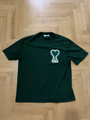 Mörkgrön t-shirt från ami paris  - Snygg mörkgrön t-shirt med ett stort hjärta och bokstaven A broderat på bröstet. Klassisk passform och rund halsringning. Perfekt för dig som vill ha en enkel men unik look. Använd tiden några gånger men endå i bra skick 
