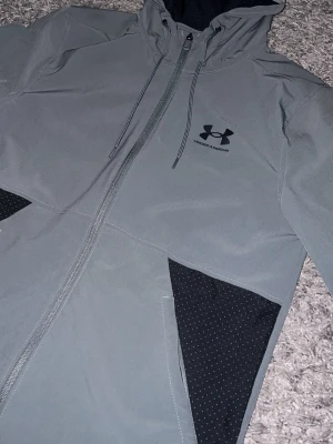  Under Armour - Snygg grå jacka från Under Armour med svart logga på bröstet. Jackan har huva med dragsko, hel dragkedja och svarta detaljer vid fickorna. Materialet är slätt och vattenavvisande, perfekt för blåsiga dagar. Priset kan diskuteras😃