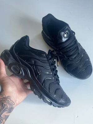  Nike Air Max Plus TN - Säljer ett par helsvarta Nike Air Max Plus sneakers med mesh och syntetiska detaljer. Skorna har den ikoniska vågiga designen, synlig Air-dämpning i sulan och svarta snören. Perfekta för dig som gillar streetwear och sportig stil.