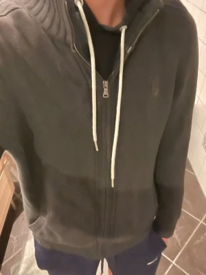 Mörkgrå stickad kofta med dragkedja - Mörkgrå stickad  med dragkedja framtill . Tröjan har ribbade muddar vid ärmslut och nederkant samt diskret emblem på bröstet. Perfekt för dig som gillar en avslappnad och stilren look.