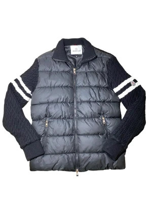 Svart pufferjacka - Snygg svart pufferjacka med stickade ärmar och vita ränder på ärmarna. Jackan har dragkedja framtill, två fickor med dragkedja och Moncler-logga på ena ärmen. Kroppen är quiltad och vadderad medan ärmarna är i grovstickad ullmix. INTE ÄKTA!
