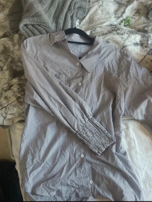 💙 H&M blårandig blus med smock | M/L | Skjortmodell - Säljer en superfin blårandig blus från H&M i storlek M/L. Den har en klassisk skjortdesign med fina blå ränder och en smockdetalj längst ner som ger en snygg och avslappnad passform. Perfekt att bära till jeans, shorts eller vita linnebyxor.