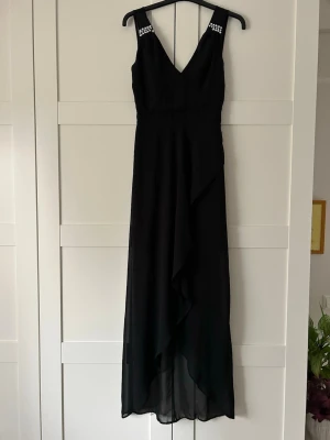 Svart långklänning med v-ringning - Elegant svart långklänning med v-ringning och smala axelband dekorerade med pärlor. Klänningen har omlottliknande fall framtill och är tillverkad i ett lätt, flödande material som ger en snygg siluett. OBS! Saknas 2 st stenar på ena sidan som syns på bilderna. Annars bra skick och lite använd.