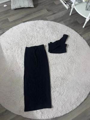 Svart ribbat set med kjol och topp - Snyggt svart ribbat set med lång kjol och matchande crop top. Kjolen är hellång och toppen är axelbandslös, vilket ger en trendig look. Perfekt för dig som gillar stilrena och enkla plagg med en modern twist.