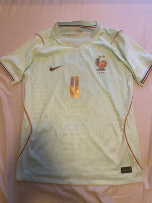 Nike Frankrike Player Edition – Michael Olise – Limited Edition - Ljusgrön fotbollströja från Nike med Frankrikes landslagsemblem och nummer 11 i guld på både fram- och baksida. Namnet 'OLISE' står på ryggen. Tröjan har korta ärmar med randiga muddar och tunna orange detaljer längs sidorna. Tillverkad i lätt och ventilerande material.