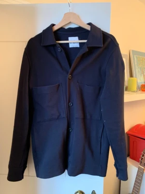 Mörkblå overshirt - Stilren mörkblå overshirt med klassisk krage och stora utanpåliggande bröstfickor. Modellen har knäppning framtill med tre knappar och långa ärmar med knapp vid ärmslut. Materialet ser ut att vara mjukt och lite tjockare, perfekt för lager på lager.