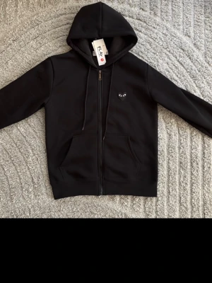 Comme de garcon kofta - Säljer en svart hoodie med dragkedja och huva. På bröstet finns ett litet svart hjärta med ögon och på ryggen ett större matchande hjärta. Hoodien har fickor framtill och ribbade muddar. Perfekt för dig som gillar stilrena plagg med en twist.