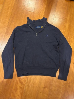 Mörkblå stickad tröja Polo Ralph Lauren - Mörkblå stickad tröja från Polo Ralph Lauren med halv dragkedja och diskret broderad logga på bröstet. Tröjan har ribbade muddar vid ärmslut och nederkant samt hög krage. Perfekt för lager på lager och klassisk stil.