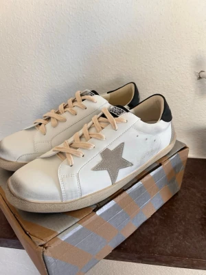 Vita sneakers med beige stjärna - Snygga vita sneakers med beige stjärndetalj på sidan och svarta detaljer vid hälen. Skorna har beige snören och en beige sula. Tillverkade i läder med mockadetaljer för en trendig look. Perfekta för dig som vill ha en clean men ändå unik stil.