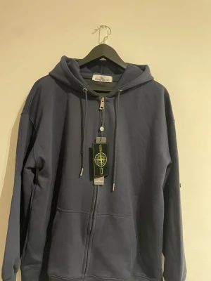 Mörkblå hoodie från Stone Island - Snygg mörkblå hoodie från Stone Island med dragkedja framtill, huva med dragsko och klassisk logotyp på taggen. Tillverkad i mjukt bomullsmaterial och har två fickor framtill. Perfekt för en avslappnad och trendig stil.                        🚨Storlek (M) passar också Storkek (S)🚨.                Pris Kan Diskuteras 🙌