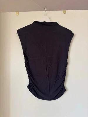 Mörkgrå ärmlös topp med rynkade sidor - Snygg mörkgrå ärmlös topp med rynkade detaljer längs sidorna som ger en cool och smickrande passform. Tillverkad i ett mjukt och stretchigt material som sitter skönt mot kroppen. Perfekt att styla med jeans eller kjol för en trendig look.