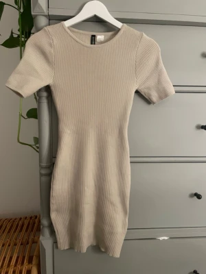 Beige ribbad tajt klänning  - Säljer en beige ribbad tajt  klänning med rund halsringning och korta ärmar. Klänningen har en slim passform och är tillverkad i ett stretchigt material som sitter snyggt på kroppen. Perfekt för en enkel och stilren look.