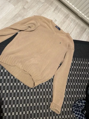 Beige stickad tröja från Polo Ralph Lauren - Snygg beige stickad tröja från Polo Ralph Lauren med rund halsringning och diskret logga broderad på bröstet. Tröjan har långa ärmar och ribbade muddar vid ärmslut och nederkant. Perfekt för lager på lager och enkel att matcha. Can negotiate for lower price
