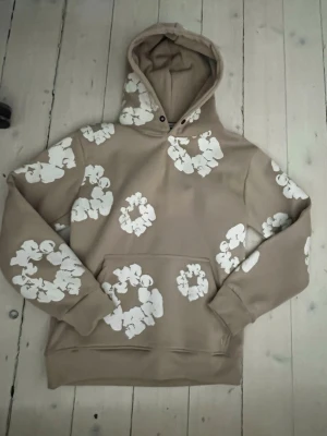 Beige hoodie med vita blommor - Snygg beige hoodie från FLEX-STYLE med vita blommönster över hela tröjan. Hoodien har en klassisk huva och magficka framtill. Tillverkad i mjukt material som känns skönt mot huden. Perfekt för dig som vill sticka ut med en unik stil.
