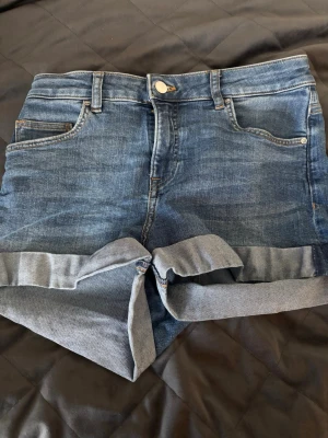Shorts  - Snygga blå denimshorts med klassisk femficksdesign och uppvikta benslut. Shortsen har hög midja och stängs med knapp och dragkedja. Perfekta för varma dagar och enkla att matcha med olika toppar.