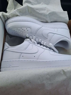 Vita Nike Air Force 1 sneakers - Klassiska helvita Nike Air Force 1 sneakers med låg profil. Skorna har en stilren design med perforerad tå, vita skosnören och den ikoniska swoosh-loggan på sidan. Tillverkade i läder med en robust gummisula för extra komfort och streetkänsla.