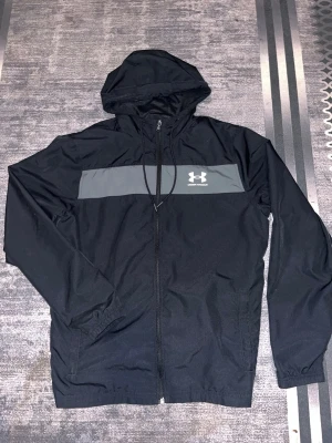 Svart vindjacka från Under Armour - Snygg svart vindjacka från Under Armour med huva och dragkedja framtill. Jackan har en grå panel över bröstet med logga, justerbar huva och meshfoder för extra komfort. Perfekt för blåsiga dagar och sportiga outfits.