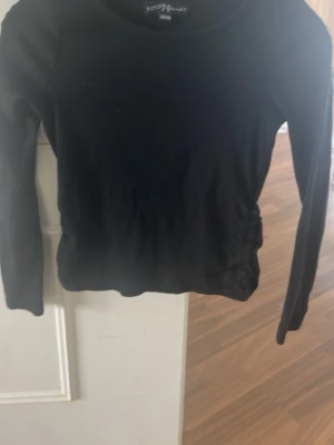 Svart långärmad topp med ribbade detaljer - Snygg svart långärmad topp med ribbade detaljer på ärmarna och en tight passform. Rund halsringning och dekorativ knytning i sidan ger en cool touch. Perfekt att matcha med jeans eller kjol för en stilren look.