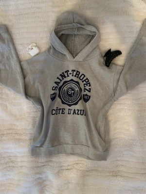 Grå hoodie Saint-Tropez TENKA - Grå hoodie från TENKA med tryck 'Saint-Tropez Côte d'Azur' i mörkblått framtill. Klassisk huva och långärmad modell i mjukt material, perfekt för en avslappnad look. Passar dig som gillar enkel och stilren streetwear.