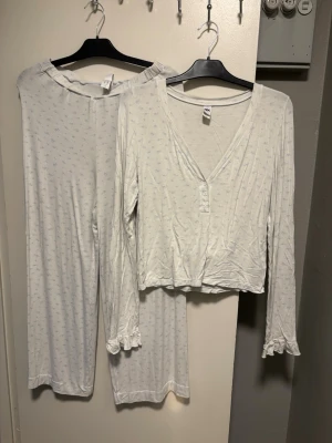 Vit pyjamas med blått mönster mk M - Mysig tvådelad pyjamas från mk i vitt med små ljusblå mönster. Setet består av långärmad topp med knappar framtill och volangdetaljer vid ärmslut samt matchande byxor med lös passform. Perfekt för chillkvällar hemma. S byxa M tröja