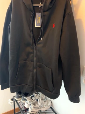 Svart hoodie från Polo Ralph Lauren med lappen kvar. - Snygg svart hoodie från Polo Ralph Lauren med dragkedja framtill och klassisk röd logga broderad på bröstet. Hoodien har huva med dragsko och stora fickor framtill. Perfekt för en chill och stilren look. Lite stor i storleken. 