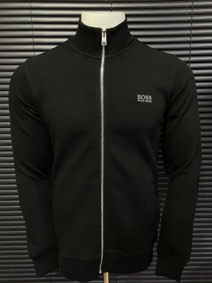 Svart och vit zip hoodie från Hugo Boss - Två stilrena hoodies från Hugo Boss, en i svart och en i vitt. Båda har hel dragkedja framtill, hög krage och diskret logga på bröstet. Tillverkade i mjukt material med ribbade muddar vid ärmslut och nederkant. Perfekt för en clean och modern look. Finns både S och M storlek 