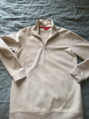 Beige halvzip tröja från RYVLS - Snygg beige halvzip tröja från RYVLS med hög krage och dragkedja framtill. Tillverkad i mjukt material och har ribbade muddar vid ärmslut och nederkant. Perfekt för en avslappnad och stilren look.