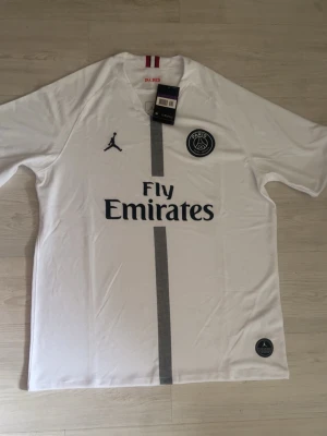 Vit PSG matchtröja Jordan x Fly Emirates - Säljer en vit PSG matchtröja med Fly Emirates-tryck och Jordan-logga. Tröjan har en grå vertikal rand i mitten, rund hals och korta ärmar. Materialet är lätt och andas, perfekt för fotboll eller träning. PSG-emblemet sitter på bröstet.