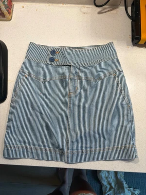 Randig jeanskjol Low waist - Snygg kort jeanskjol från Maine New England med blåvita ränder och två blå knappar i midjan. Kjolen har klassiska fickor fram och är gjord i ett slitstarkt bomullsmaterial. Perfekt för dig som gillar en avslappnad och trendig stil.