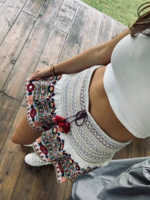 Broderad vit kjol med tofsar - Vit kjol med färgglad brodyr i rött, gult, blått och rosa. Hög midja med smock och dekorativa tofsar framtill. Blommönster och volangkant ger en bohemisk vibe. Perfekta för sommaren och festivaler.
